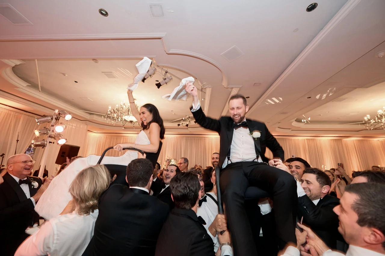 Alex and Jamie laughing as their guests lift them on chairs during the hora at their wedding reception