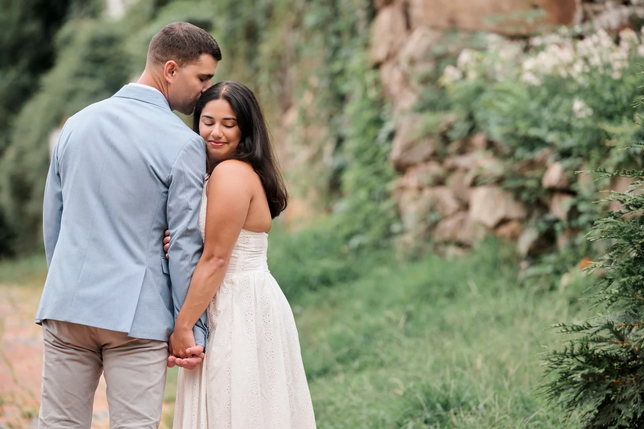 Eric kisses Anjali’s forehead as they hold hands beside a leafy stone wall during their Occoquan engagement session