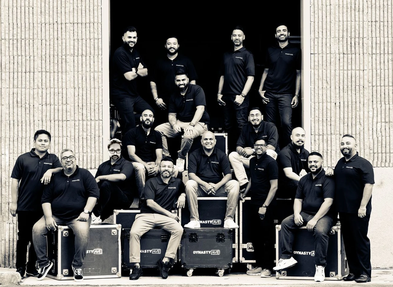The Dynasty AVE team posing by an open doorway, wearing Dynasty AVE shirts and seated or standing around audio equipment cases