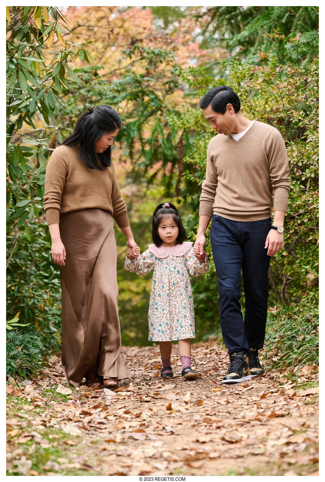 Two parents holding their daughter's hands on an autumn path at McCrillis House and Gardens, photographed by The Regeti’s