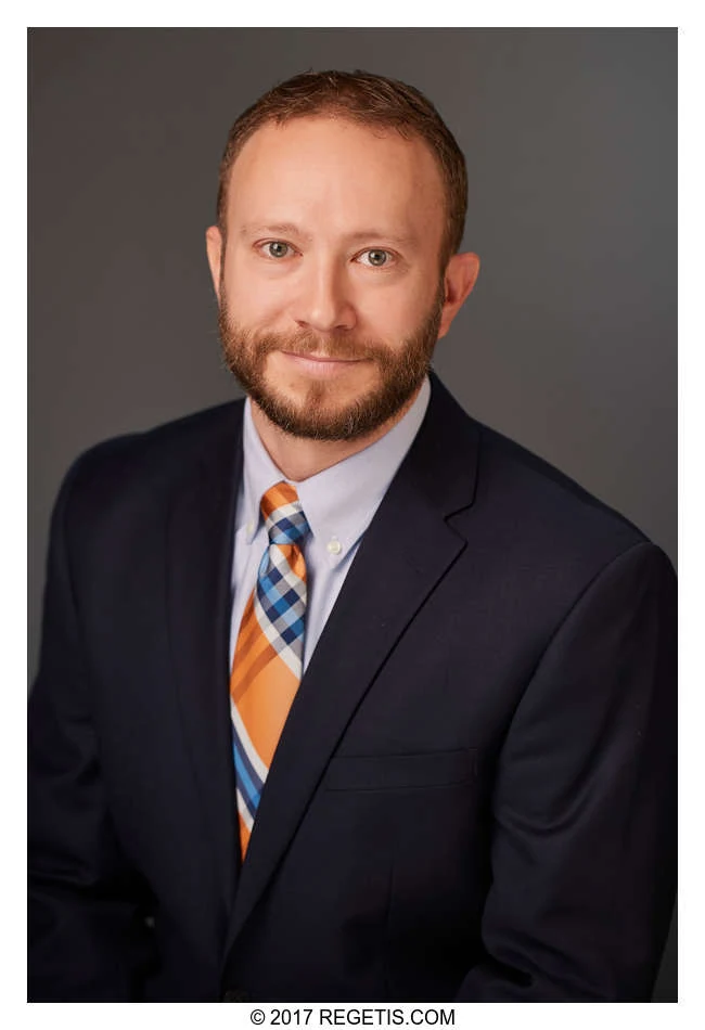 A professional in a navy suit and striped tie, photographed against a charcoal background by The Regeti’s