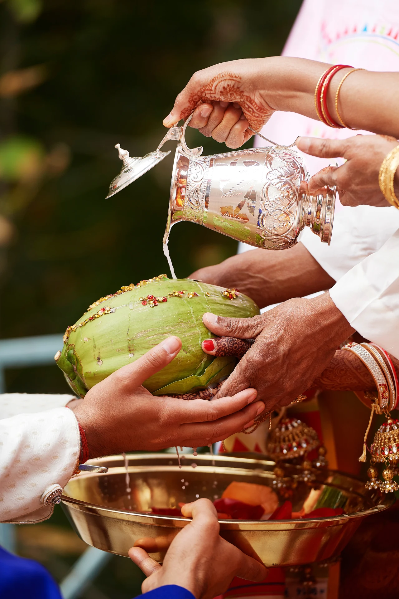 Family hands pour water from a silver vessel over a decorated coconut during Kanya Daan.